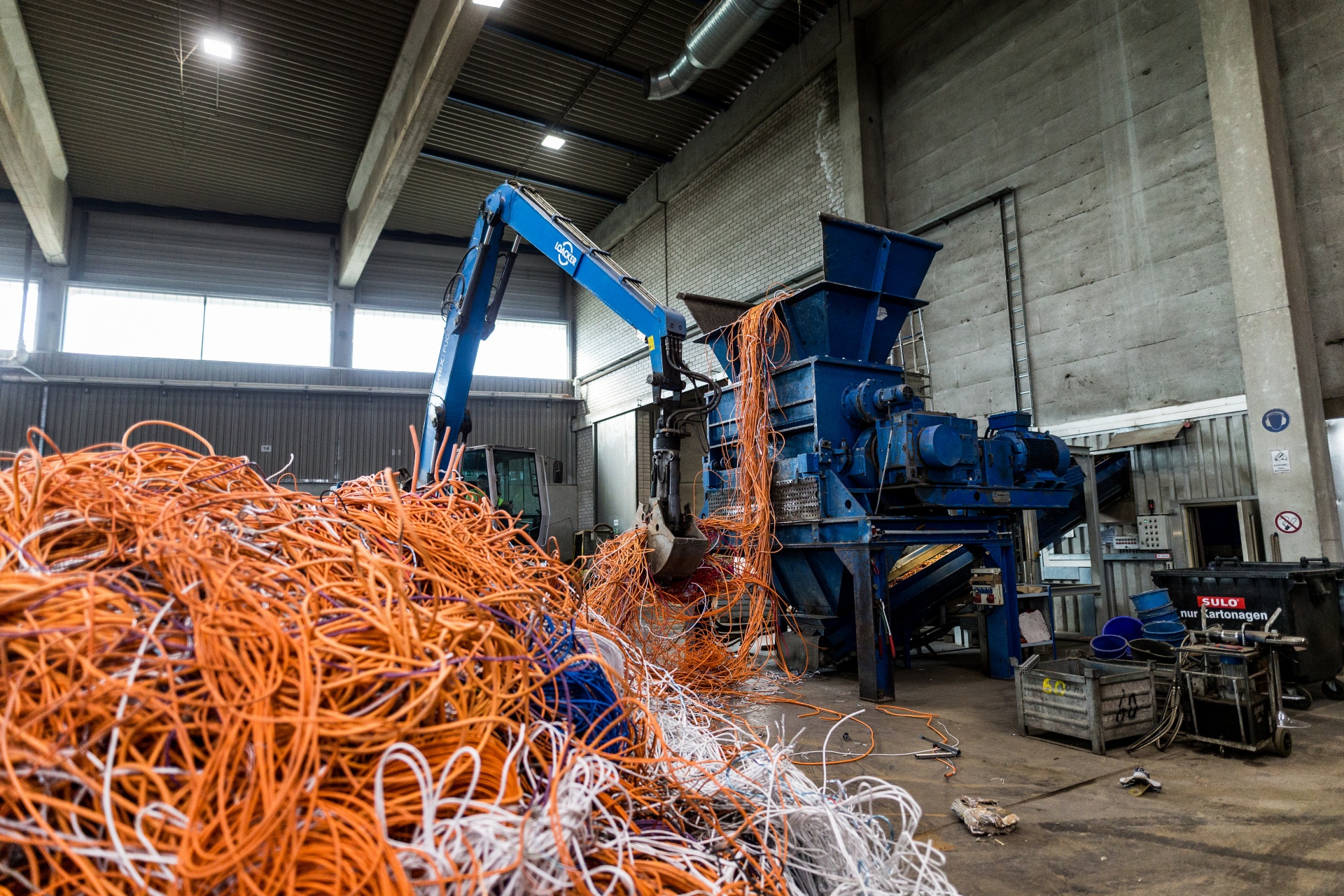 Eine blaue Maschine, die einen Haufen Kabel greift und in einen blauen Schredder transportiert. Im Vordergrund ist ein großer Haufen von Kabel sind orange und weiß. Wir befinden uns in einer Produktionshalle.
