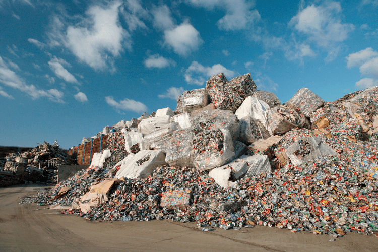 Ein großer Müllberg aus gepressten und losen Aluminiumdosen sowie Plastikflaschen auf einem Recyclinghof unter blauem Himmel mit weißen Wolken. Die gepressten Ballen sind teilweise in weißen Plastikhüllen verpackt.