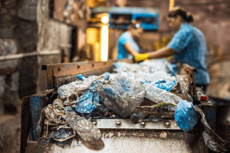 Nahaufnahme eines Förderbands mit verschmutzten Plastikflaschen und Kunststoffabfällen in einer Recyclinganlage. Im Hintergrund sortieren zwei Arbeiterinnen in blauen Hemden und mit gelben Handschuhen den Müll.