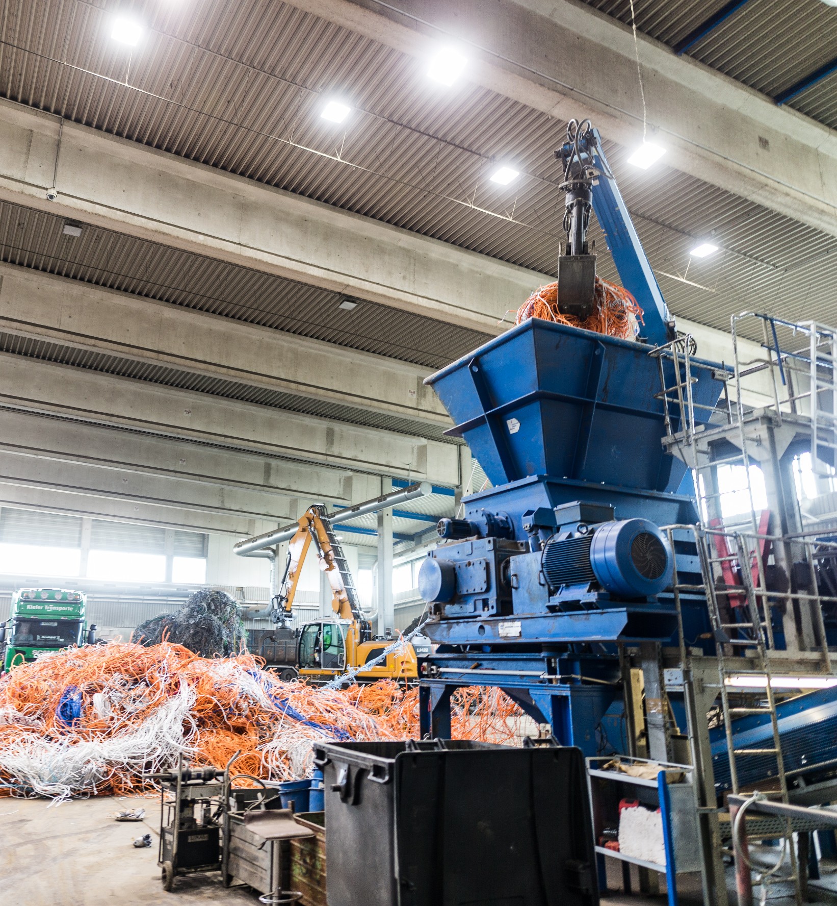 Eine blaue Maschine, die einen Haufen Kabel greift und in einen blauen Schredder transportiert. Die Kabel sind orange und weiß. Wir befinden uns in einer Produktionshalle.